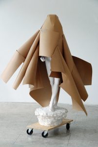 Weiße Gipsfigur mit einem Mantel aus Packpapier auf einem Rollbrett